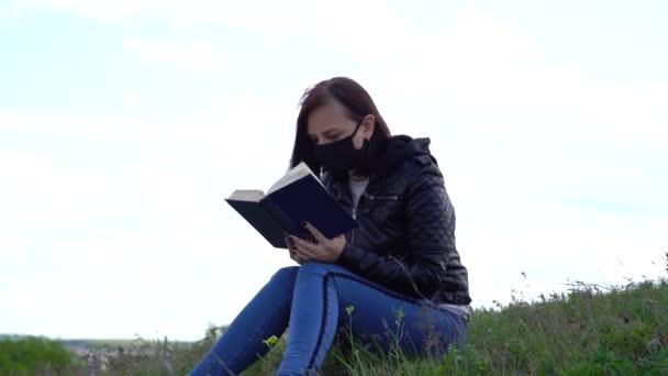 Primo piano di giovane donna in maschera medica nera e vestiti casual seduto sulla collina e la lettura di libro il giorno nuvoloso. Donna adulta che riposa sola in campagna. — Video Stock