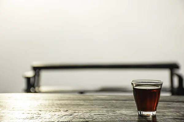 Un vaso de café negro sobre una mesa de madera Niebla de fondo y luz del sol de la mañana . — Foto de Stock