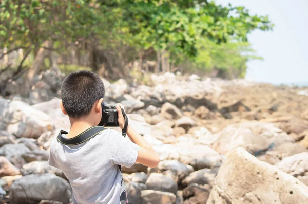 The image behind Asian boy holding a camera to take pictures Background blurry rocks and tree at Laem Kho Kwang Beach  in Chumphon , Thailand. March 18, 2020
