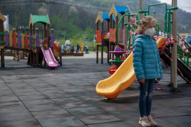 Küçük bir kız, tıbbi maske takıyor, arka planda oyun parkı kapalı. Koruyucu maskeli bir çocuk. COVID-19 'a karşı tehlike ve güvenlik önlemleri uyarısı. Koronavirüs salgını
