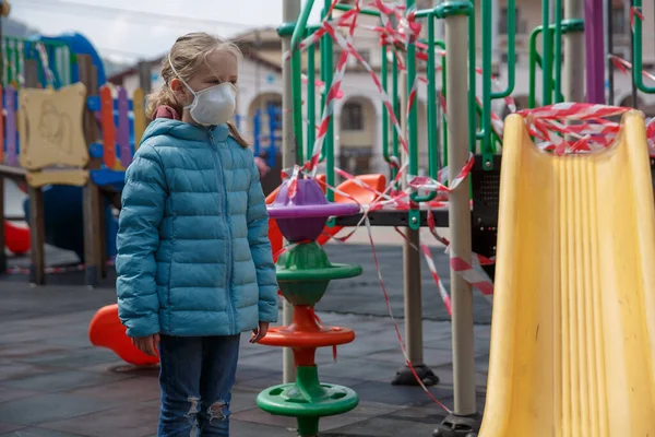 Küçük bir kız, tıbbi maske takıyor, arka planda oyun parkı kapalı. Koruyucu maskeli bir çocuk. COVID-19 'a karşı tehlike ve güvenlik önlemleri uyarısı. Koronavirüs salgını