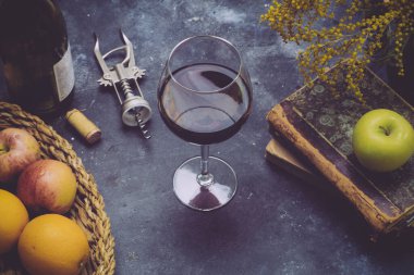 Koyu arkaplanda bir kadeh kırmızı şarap. Masada meyve, şişe ve eski kitaplarla birlikte şarap var. Rahatla ve romantizm kavramı.