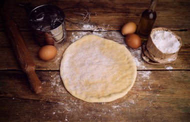 Ev mutfağında pizza hamuru pişiyor. Ekmek, pizza, börek ve ekmek için ev yapımı hamur işi. Ahşap bir arka planda hamur malzemeleri (un, yumurta, tereyağı).