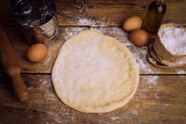 Ev mutfağında pizza hamuru pişiyor. Ekmek, pizza, börek ve ekmek için ev yapımı hamur işi. Ahşap bir arka planda hamur malzemeleri (un, yumurta, tereyağı).