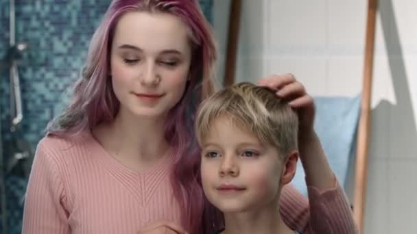 Chica joven está peinando el pelo de su hermano pequeño delante de un espejo — Vídeo de stock