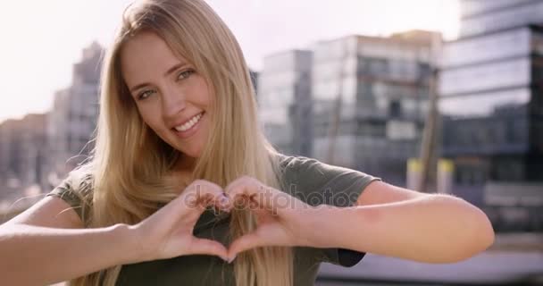 Attractive blonde woman making a heart with her hands infront of a modern city background — Stock Video