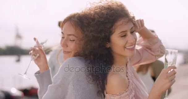 Две девушки танцуют спина к спине, с очками в руках и друзьями на заднем плане — стоковое видео