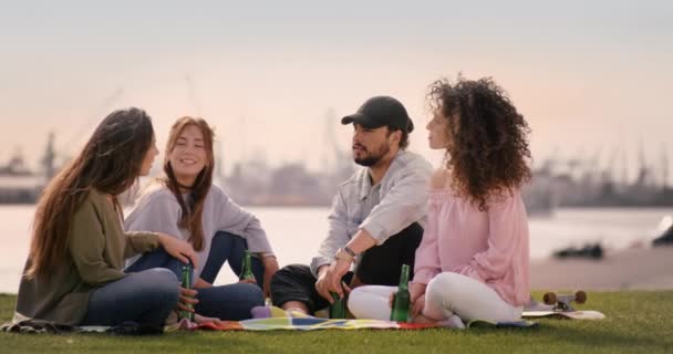 Jovens pessoas bonitas bebendo e desfrutando do tempo ensolarado enquanto se sentam em um prado — Vídeo de Stock