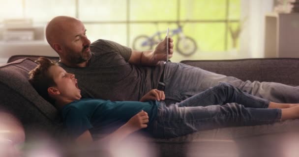 Son typing on the tablet his father is holding on the couch — Stock Video