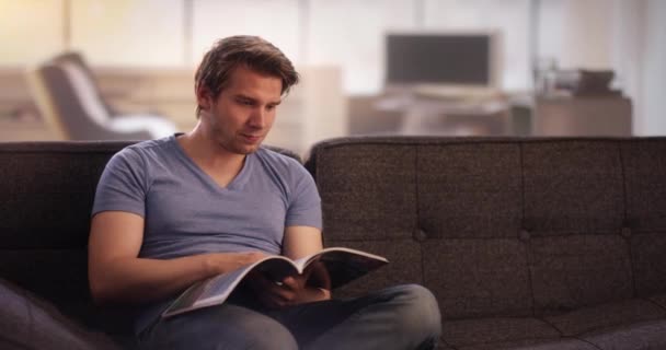 Молодой человек читает книгу на диване — стоковое видео