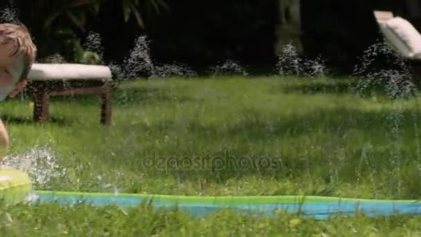 Мальчик веселится на водной горке в зеленом саду летом — стоковое видео