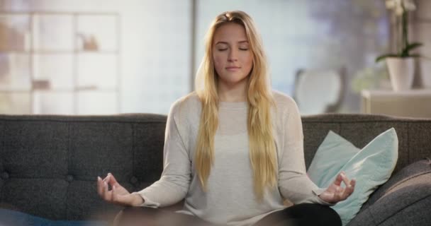Красива молода леді відкриває очі, сидячи в легкій позі йоги на дивані — стокове відео
