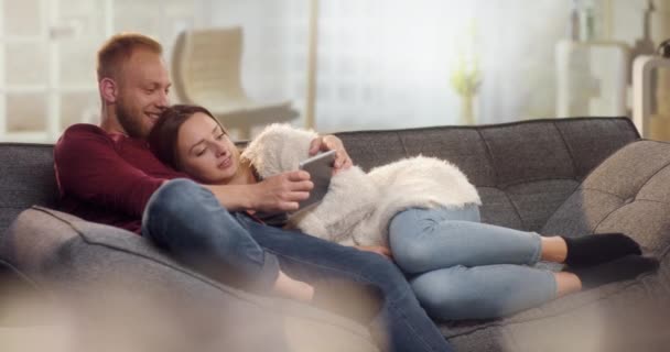 Δροσερό ζευγάρι στον καναπέ αγκαλιά και βλέποντας αστεία πράγματα στο tablet — Αρχείο Βίντεο
