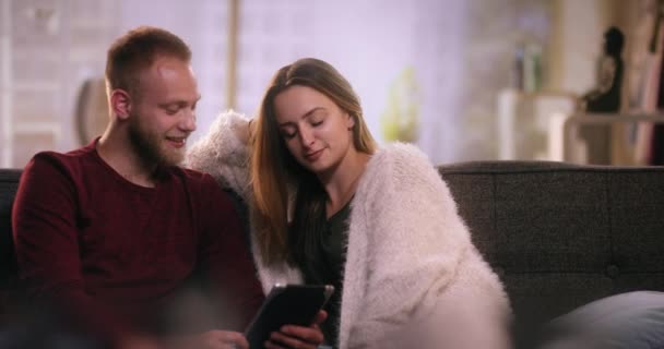 Молодая пара сидит на диване и смотрит на планшет — стоковое видео