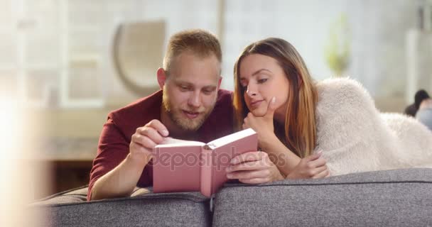 Junges süßes Paar liegt auf einer Couch und liest ein Buch — Stockvideo