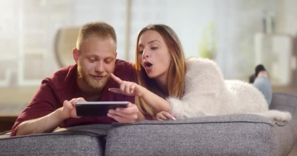 Молодая милая пара лежит на диване и делает что-то на планшете — стоковое видео