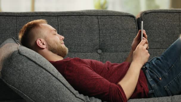 英俊的年轻男子，躺在沙发上读有趣的消息，在平板电脑上 — 图库视频影像