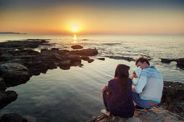Um jovem casal com um bebé na praia ao pôr-do-sol. Família — Fotografia de Stock