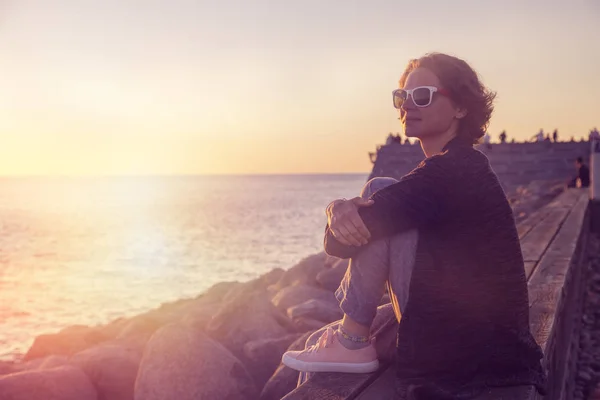 夕日を見ながらビーチに座っている美しい若い女性 — ストック写真