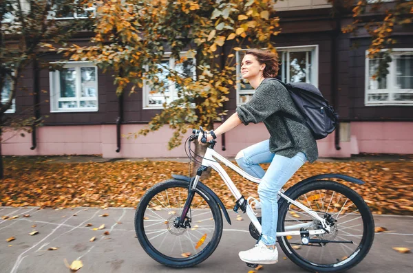 Hermosa joven estudiante en un suéter y jeans en una bicicleta — Foto de Stock