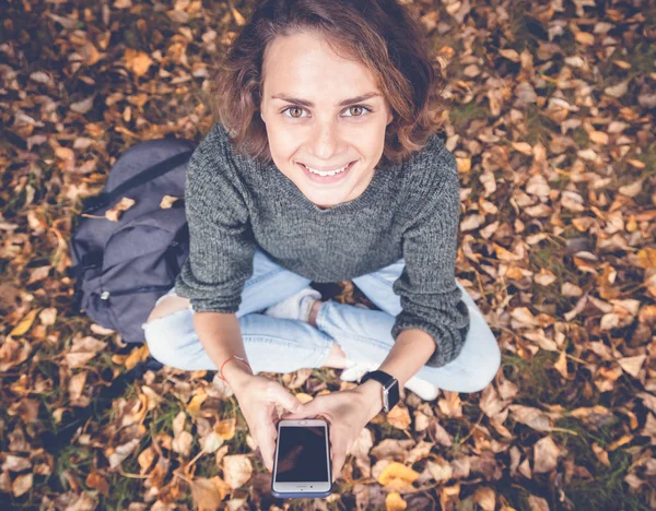 Молодая счастливая красивая женщина сидит на осенней листве с мобильным p — стоковое фото