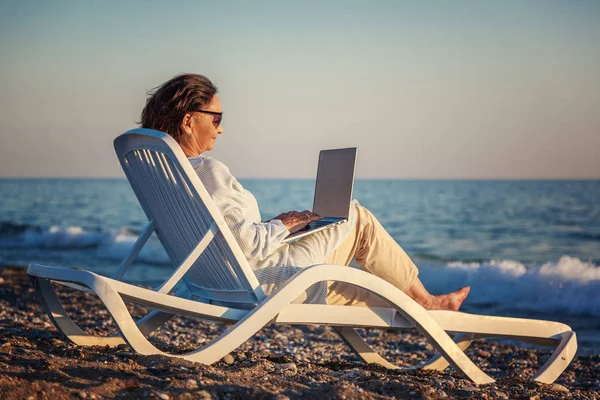 Stijlvolle aantrekkelijke rijpe vrouw 50-60 maakt gebruik van de laptop op de kust, reizen en pensioen, mode en schoonheid, actieve ouderen — Stockfoto