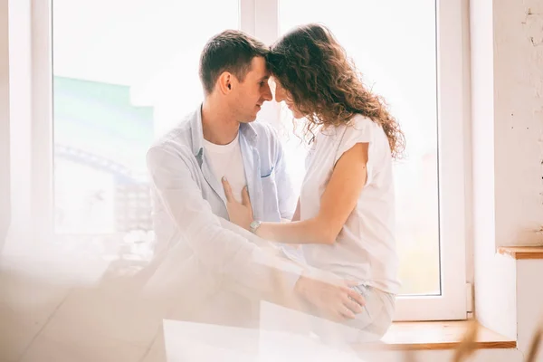 Joven pareja feliz abrazándose en el alféizar de la ventana en su apartamento , — Foto de Stock