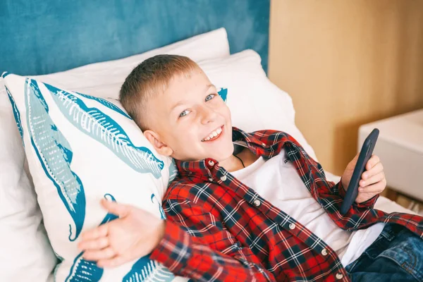 Feliz niño alegre 8 años colegial se encuentra en la cama con el móvil —  Fotos de Stock