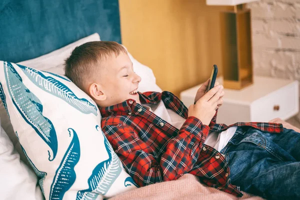 Feliz niño alegre 8 años colegial se encuentra en la cama con el móvil —  Fotos de Stock