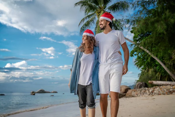 Noel Baba şapkalı genç çift kıyı boyunca yürüyor. — Stok fotoğraf
