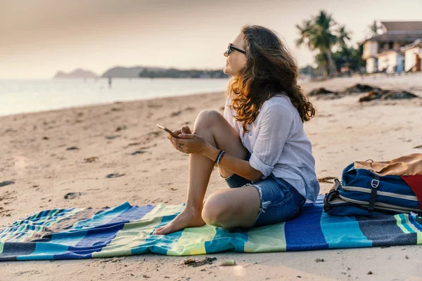 Junge schöne Frau sitzt mit einem Smartphone im Sand eines Strandes — Stockfoto