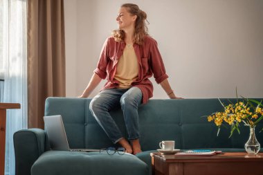 Mutlu, genç, güzel ve çekici bir kız evdeki oturma odasındaki kanepede oturuyor, işten dinleniyor ve dizüstü bilgisayarını kullanıyor..