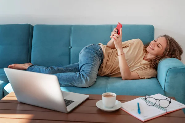 Smiling woman chatting lying on a blue sofa on smartphone. Young woman working from home, drinking coffee