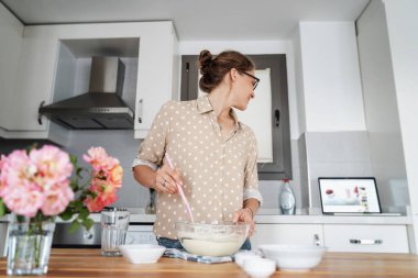 Mutfakta tatlı hazırlayan ve internette tarifi izleyen genç bir kadın, evde yemek pişirme, hobiler ve eğlence, üst manzara, hamuru mutfak spatulasıyla karıştırma.