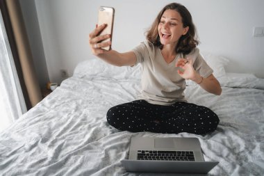 Yatakta mutlu genç bir kadın online görüntülü konuşma yapıyor, uzun çalışma eğitimi evde kalma konsepti, akıllı telefon ve dizüstü bilgisayarla yatak odasında oturuyor.