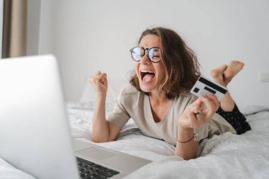 Gözlüklü mutlu genç kıvırcık kadın elinde dizüstü bilgisayar, uzaktan çalışma ve yaratıcılık, çevrimiçi yaşam, online tüketim, dijital ekonomi konseptiyle internette alışveriş yapıyor..