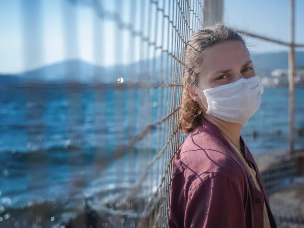 Genç bir kız tıbbi maskeli bir kadın demir ızgaradan denize bakıyor. Denize seyahat yasağı, kapalı sınır konsepti.