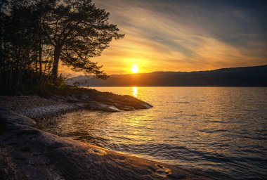 Parlak güzel manzara, Norveç 'te Snasa Gölü' nde gün batımı. Taş sahili ve ağaç silueti.