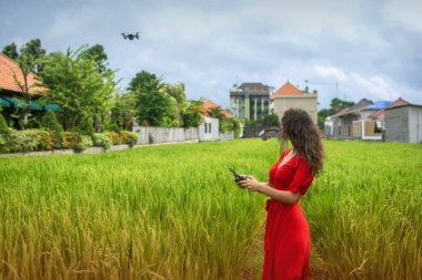 İnsansız hava aracıyla yürü. Esmer kıvırcık kız pirinç tarlasında uzaktan kumandayla İHA 'yı çalıştırıyor. Moda kırmızısı elbiseli genç bir kadın alçak uçan bir dron. Alçaktan uçan bir İHA 'ya uzanıyor.