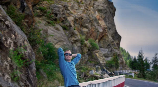 Joven en medio de la carretera y mirando hacia otro lado, disfrutando del aire fresco cerca de la hermosa montaña. Retrato de viajero con gafas de sol caminando por el camino . — Foto de Stock