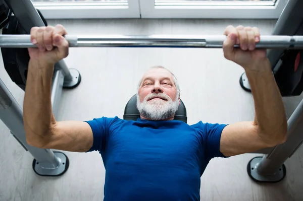 Homme âgé dans la salle de gym travaillant avec des poids, banc de pressage . — Photo