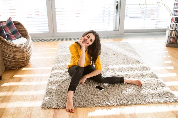 Женщина дома сидит на ковре, рядом смартфон — стоковое фото