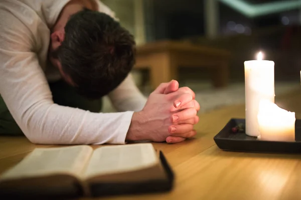 Young man praying, kneeling, Bible and candle next to him. — Stock Photo, Image