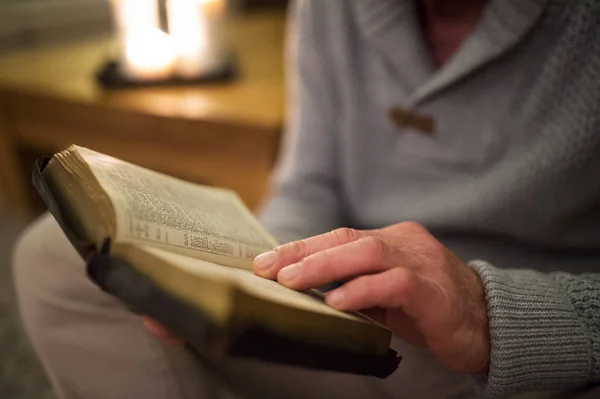 Uomo irriconoscibile a casa a leggere la Bibbia, candele accese dietro — Foto Stock