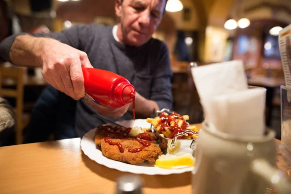 Senior man in restaurant zet ketchup op frietjes en schnitzel — Stockfoto
