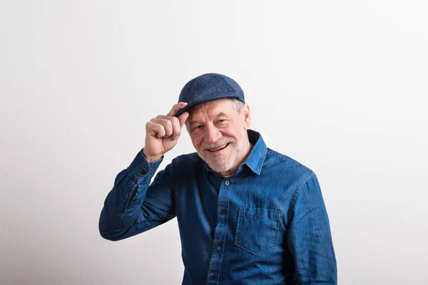 Senior man in denim shirt and flat cap smiling, studio shot. — Stock Photo, Image