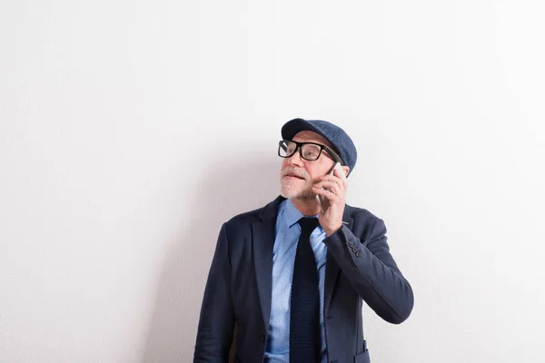 Homem sênior em camisa azul, jaqueta, óculos e boné, segurando smartphone . — Fotografia de Stock