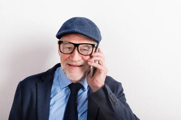 Homem sênior em camisa azul, jaqueta, óculos e boné, segurando smartphone . — Fotografia de Stock