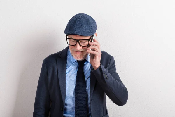 Uomo anziano in camicia blu, giacca, occhiali e berretto, in possesso di sm — Foto Stock