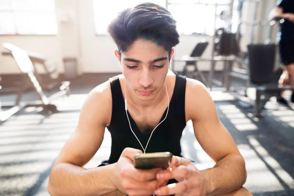 Hombre hispano en el gimnasio descansando, sosteniendo el teléfono inteligente, escuchando musi — Foto de Stock